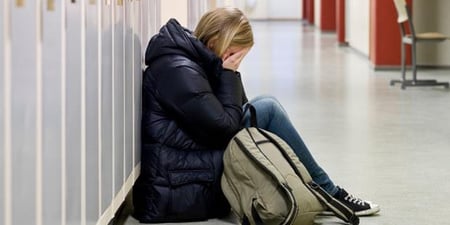 Jente sitter i skolegangen med hodet i hendene og ser ut som hun gråter. Hun har på seg boblejakke og skolesekken ligger ved siden av henne på gulvet.