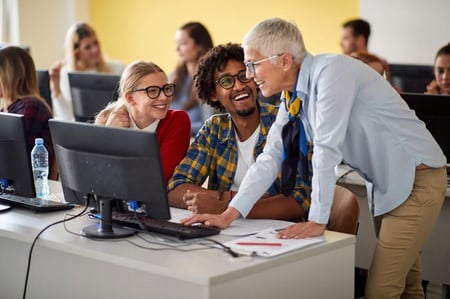1 person står bøyd for å vise 2 andre personer noe på en PC. Foto. 