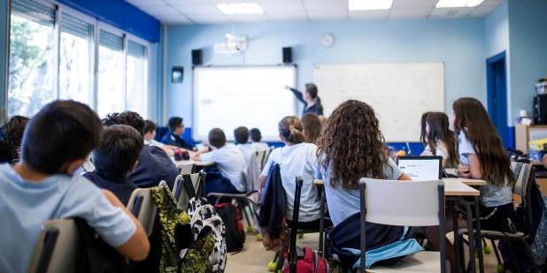 Bilde tatt bakfra i et klasserom der elevene sitter ved pultene sine og en lærer viser noe på en white board. 