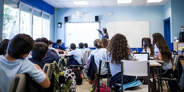 Bilde tatt bakfra i et klasserom der elevene sitter ved pultene sine og en lærer viser noe på en white board. 