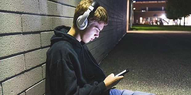 En gutt som sitter inntil en vegg. Han ser på mobiltelefonen og har hodetelefoner på. Foto.