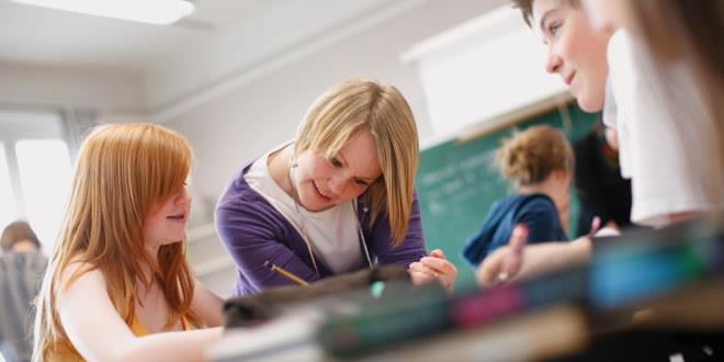 En lærer bøyer seg ned over en pult, hvor det sitter tre elever og jobber med skolearbeid.