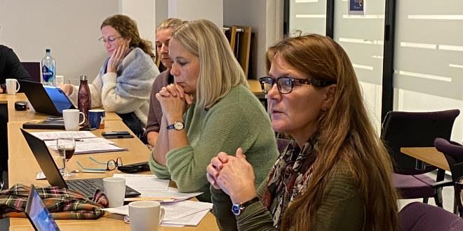 Vi ser bilde av tre personer i halvfigur. De sitte ved et bord som møtedeltakere. 