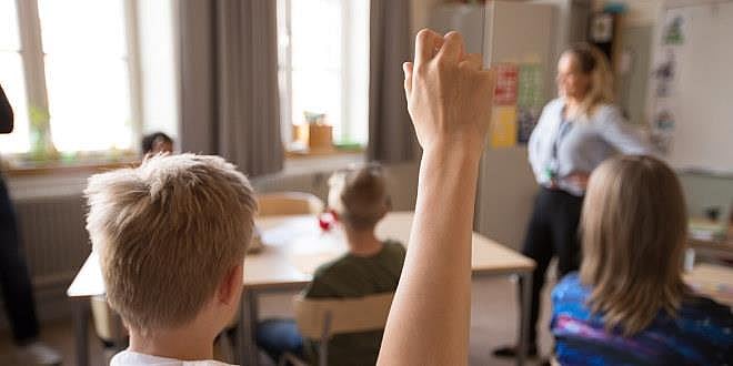 illustrasjonsfoto fra et klasserom der en gutt, sett bakfra, rekker opp hånda. 