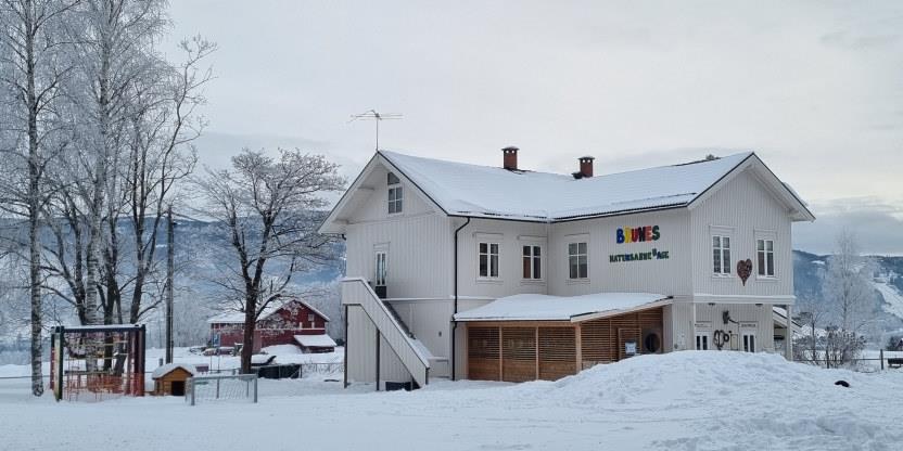 Hvitmalt barnehage, tatt utendørs, med mye snø, fjell i bakgrunnen, og noen trær og en lekeplass.