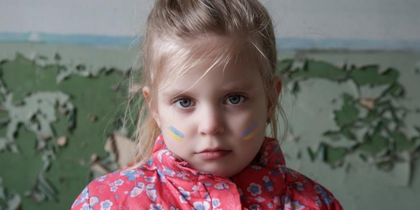 Portrettbilde av ung jente med blondt hår, rosa jakke og det ukrainske flagget malt på begge kinn mot en ødelagt murvegg der grønnmalingen flasser av.