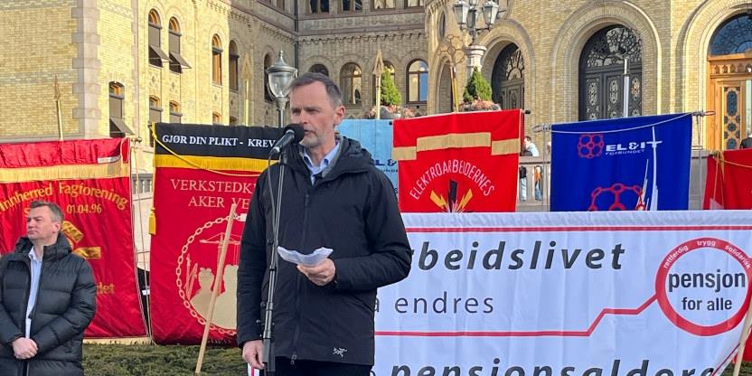 Mann i mørk jakke står foran faner ved Stortinget og taler i mikrofon.