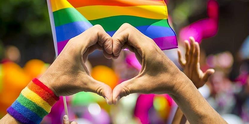 Bilde med to hender som danner et hjerte med pride armbånd og prideflagg