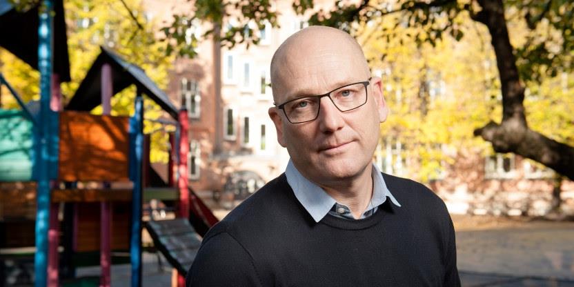 Profil av leder i Utdanningsforbundet med briller og blåskjortekrag under svart genser. Utendørs med lekeapparat og grønne trær i bakgrunnen, sol. 