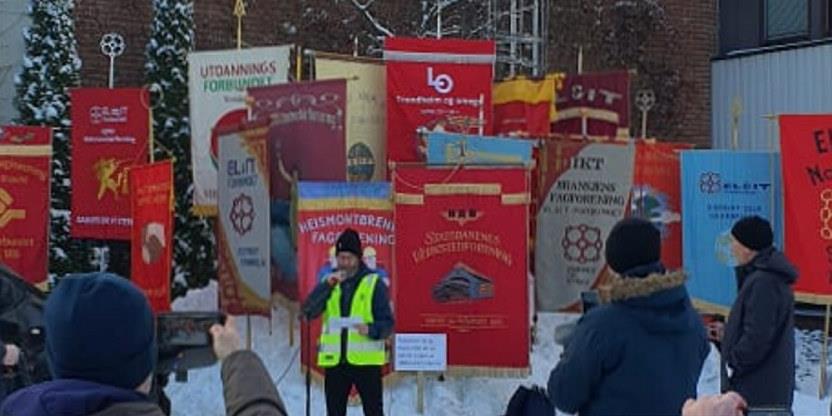 En rekke faner fra ulike fagforbund er stilt opp i snøen bak en person i refleksvest som holder en mikrofon. Folk i forgrunnen tar bilder eller filmer med mobil. Foto.