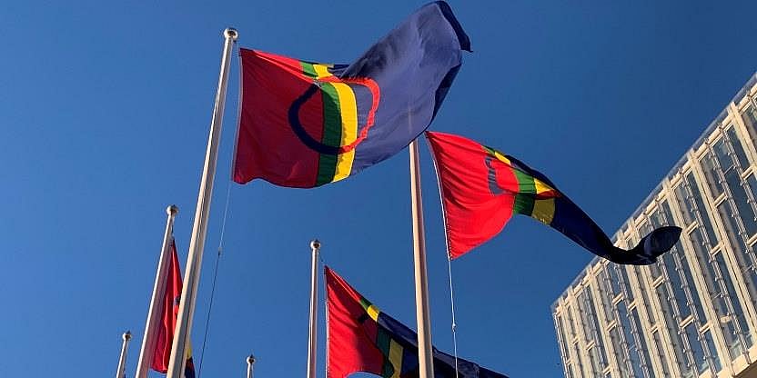 Samiske flagg vaier i vinden med blå ,skyfri himmel og et moderne bygg i glass og stål bak.