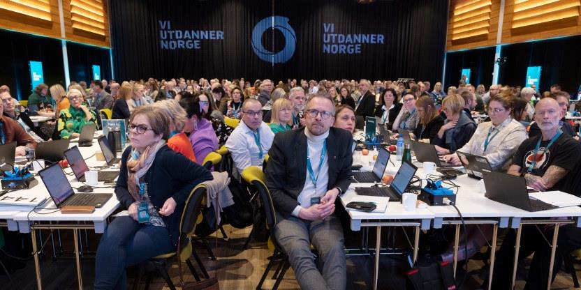 Landsmøtesalen sett forfra - stort rom med masse mennesker ved småbord, med et banner Vi Utdanner Norge i bakgrunnen.